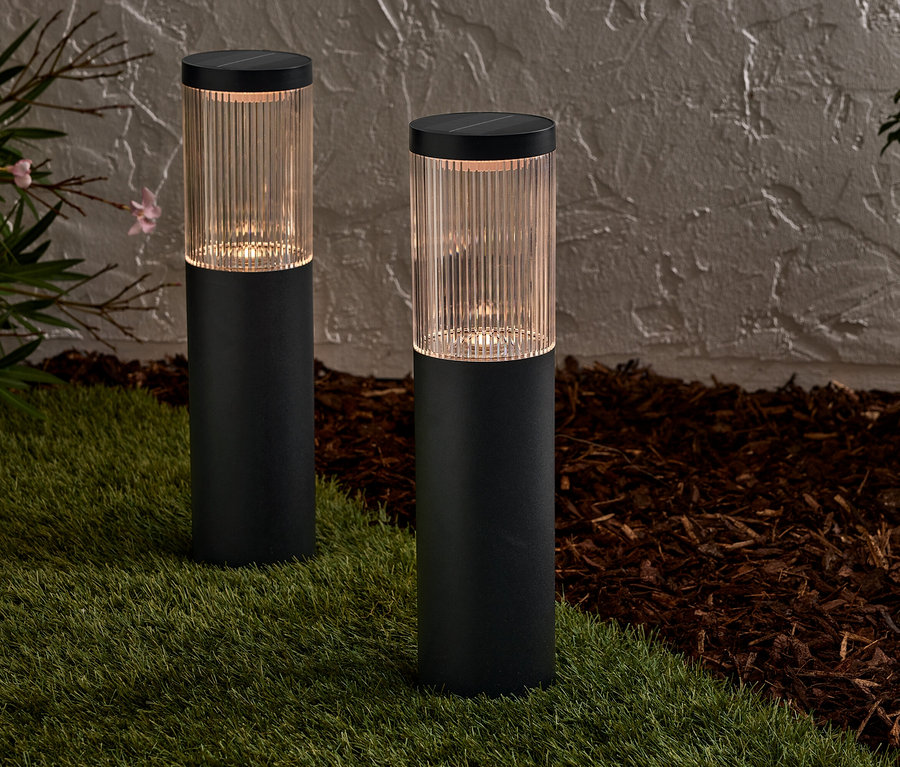 Zwei schwarze Solarlampen stehen auf einer Rasenfläche vor einer Wand.