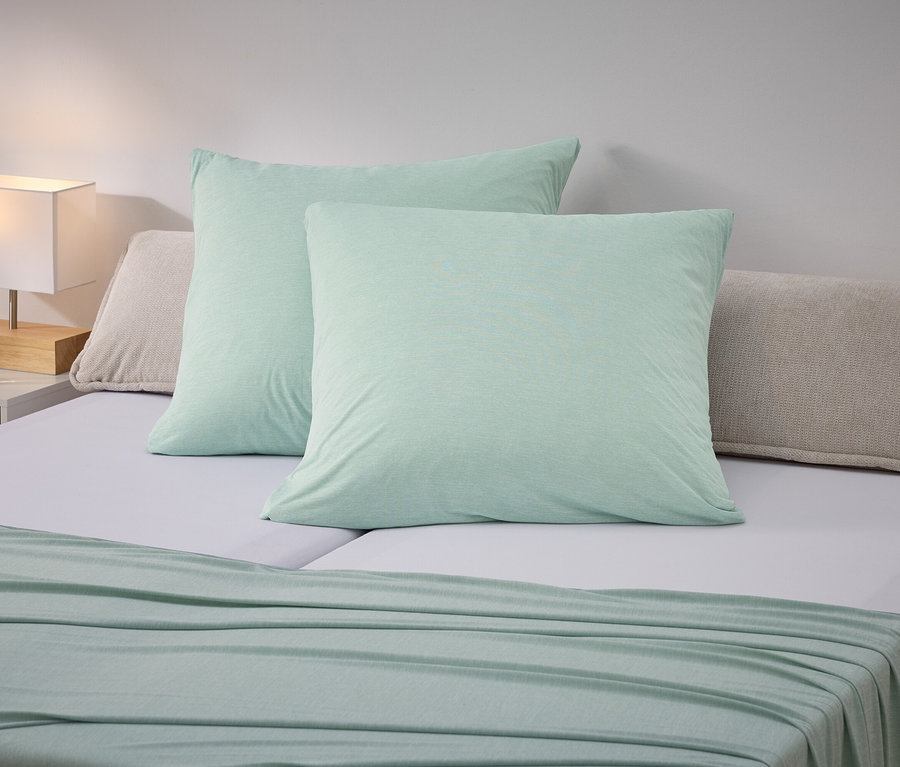 Zwei hellgrüne Kissen liegen auf einem Bett mit hellgrauem Bettlaken. Eine hellgrüne Decke liegt über das Fussende des Bettes. Ein Nachttisch mit einer Lampe steht neben dem Bett.