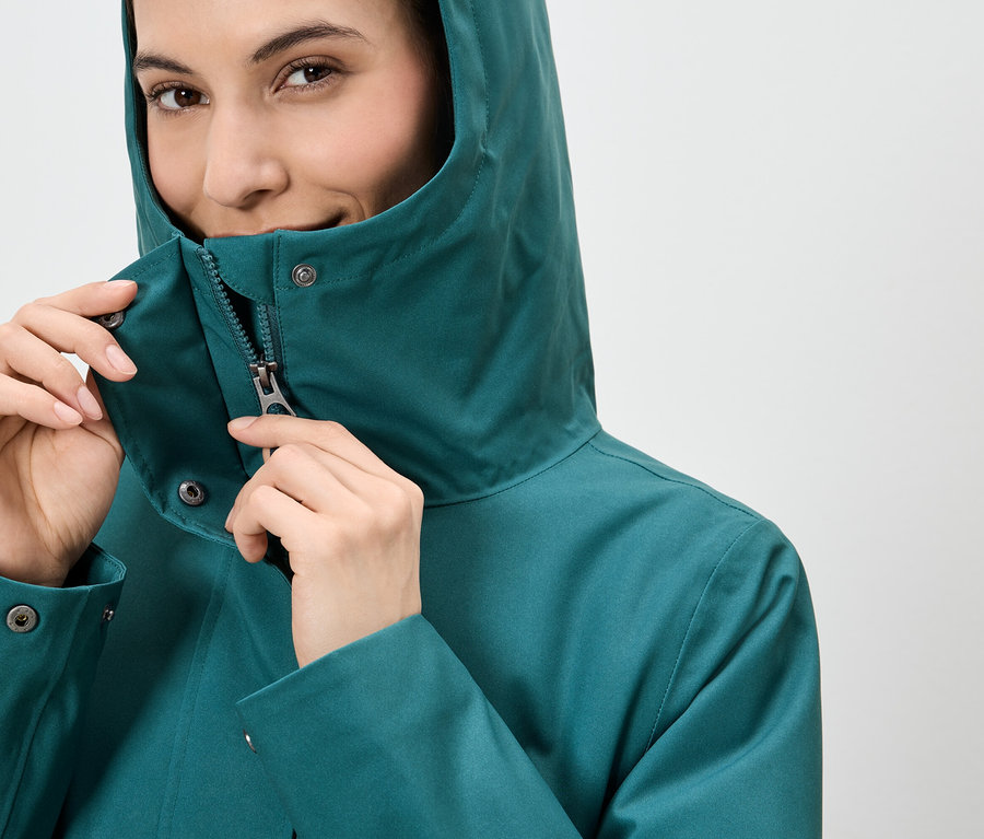 Frau schliesst den Reissverschluss einer smaragdgrünen Regenjacke mit Kapuze.
