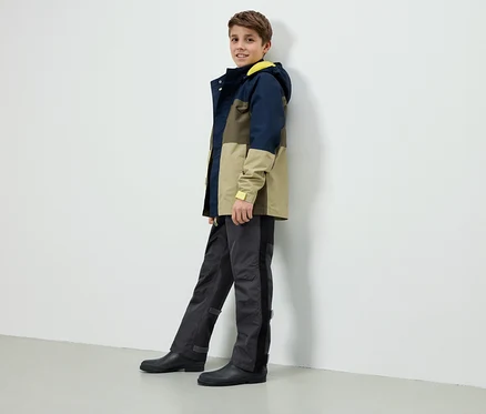 Ein Junge steht an einer weißen Wand, trägt eine farblich abgesetzte Jacke mit gelber Kapuze, graue Hosen und schwarze Gummistiefel.