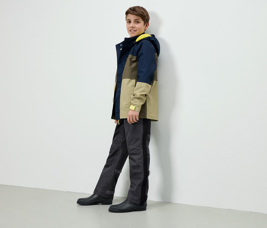 Ein Junge steht an einer weißen Wand, trägt eine farblich abgesetzte Jacke mit gelber Kapuze, graue Hosen und schwarze Gummistiefel.