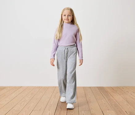 Mädchen mit langen blonden Haaren, trägt ein lila Langarmshirt, graue weite Hosen und weiße Turnschuhe.