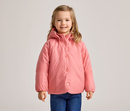 Ein junges Mädchen mit blonden Haaren, das eine rosa Kapuzen-Steppjacke und blaue Jeans trägt, lächelt in die Kamera.