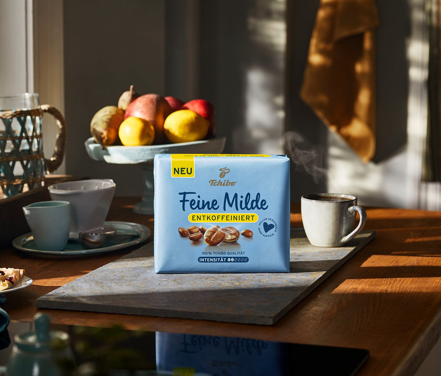 Hellblaue Tchibo Feine Milde Entkoffeiniert Kaffeepackung auf einem grauen Brett, neben einer dampfenden Tasse. Obstschale und Kuchenstück im sonnigen Küchenhintergrund.