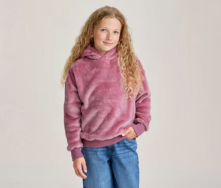 Junges Mädchen mit langen, lockigen blonden Haaren lächelt, trägt einen pinken flauschigen Kapuzenpullover und blaue Jeans, eine Hand in der Tasche.