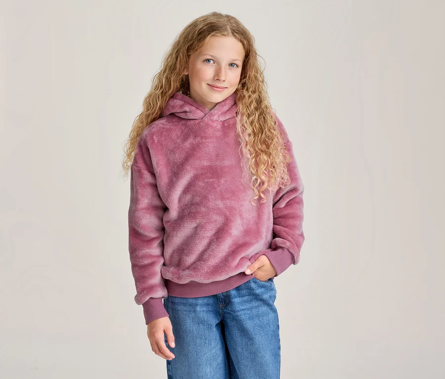 Junges Mädchen mit langen, lockigen blonden Haaren lächelt, trägt einen pinken flauschigen Kapuzenpullover und blaue Jeans, eine Hand in der Tasche.