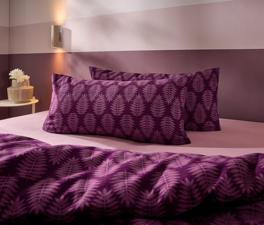Bett mit lila Bettwäsche mit Farnblattmuster, hellrosa Spannbettlaken und zweifarbiger lila Wand. Eine Wandlampe und ein Beistelltisch mit Blumenvase sind sichtbar.
