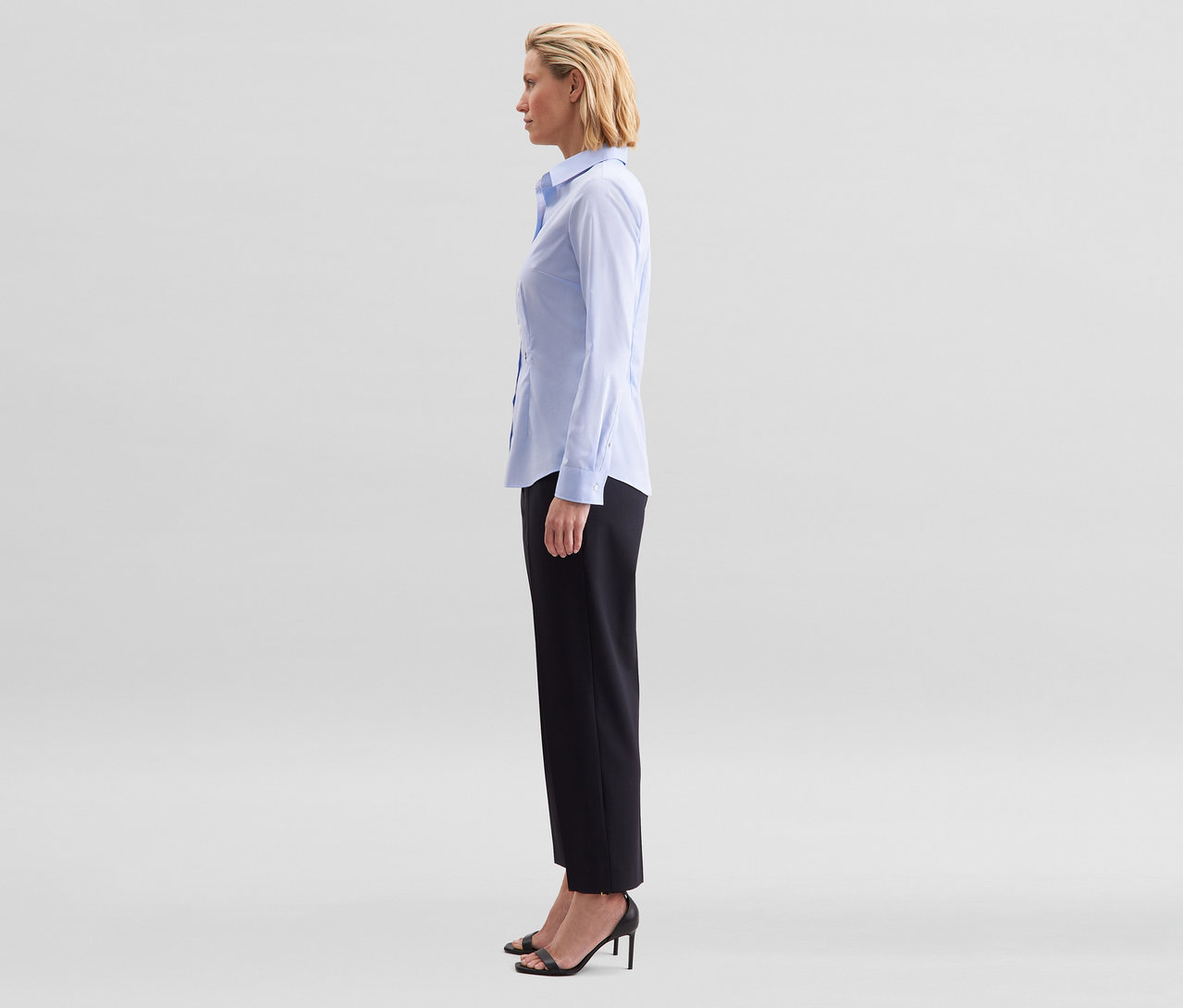 Eine Frau steht im Profil und trägt eine hellblaue Seidensticker Bluse Slim Fit non iron, schwarze Hose und schwarze Sandalen mit Absatz.