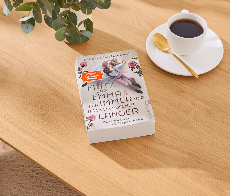 Buch „Fritz und Emma & Für Immer und Noch Ein Bisschen Länger“ von Barbara Leciejewski auf Holztisch, daneben Kaffeetasse und Pflanze.