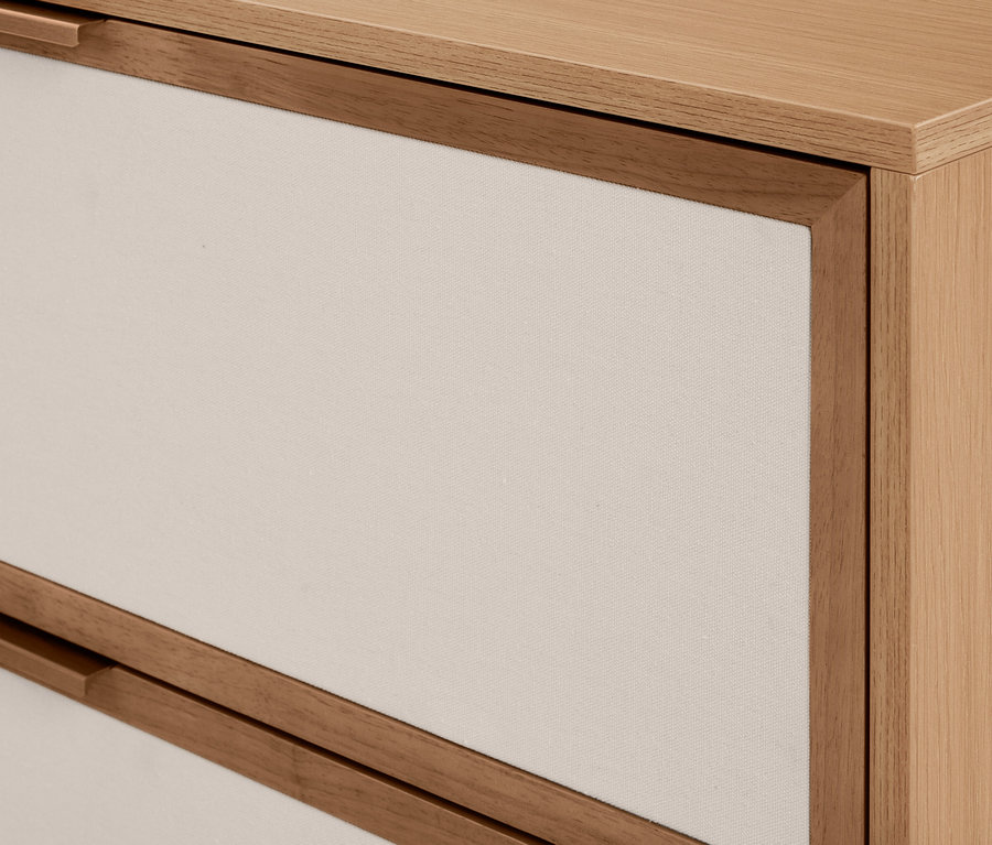 Nahaufnahme einer hellen Holzkommode mit zwei Schubladen. Die Schubladenfronten sind aus hellem cremefarbenem Stoff, von Holz gerahmt, und haben integrierte Holzgriffe.