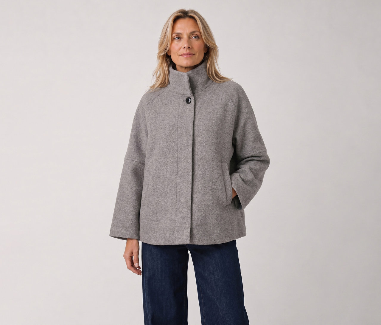 Eine blonde Frau steht in einer grauen, hochgeschlossenen Jacke mit weiten Ärmeln und dunklen Jeans, die rechte Hand in der Tasche.