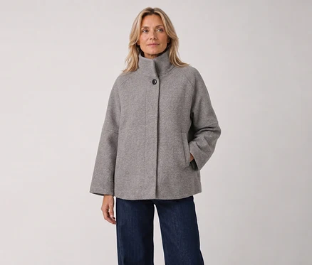 Eine blonde Frau steht in einer grauen, hochgeschlossenen Jacke mit weiten Ärmeln und dunklen Jeans, die rechte Hand in der Tasche.