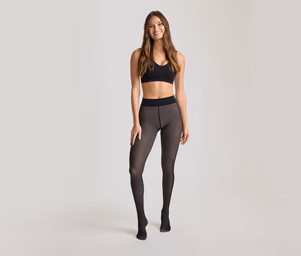 Frau steht lächelnd da und trägt einen schwarzen Sport-BH und eine transparente schwarze Strumpfhose.