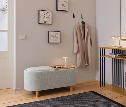 Ein gemütlicher Eingangsbereich mit einer hellgrauen Polsterbank, auf der ein Holztablett mit einer kleinen Lampe und Schlüsseln steht. Darüber hängen drei abstrakte Linienbilder und ein beiger Mantel an schwarzen Haken. Rechts davon steht eine Holzkonsole mit schwarzen Stützen, auf der zwei kleine Lampen, gefaltete Kleidung, ein Gürtel und eine geflochtene Tasche liegen. Ein dunkler gemusterter Teppich liegt auf dem Holzboden.
