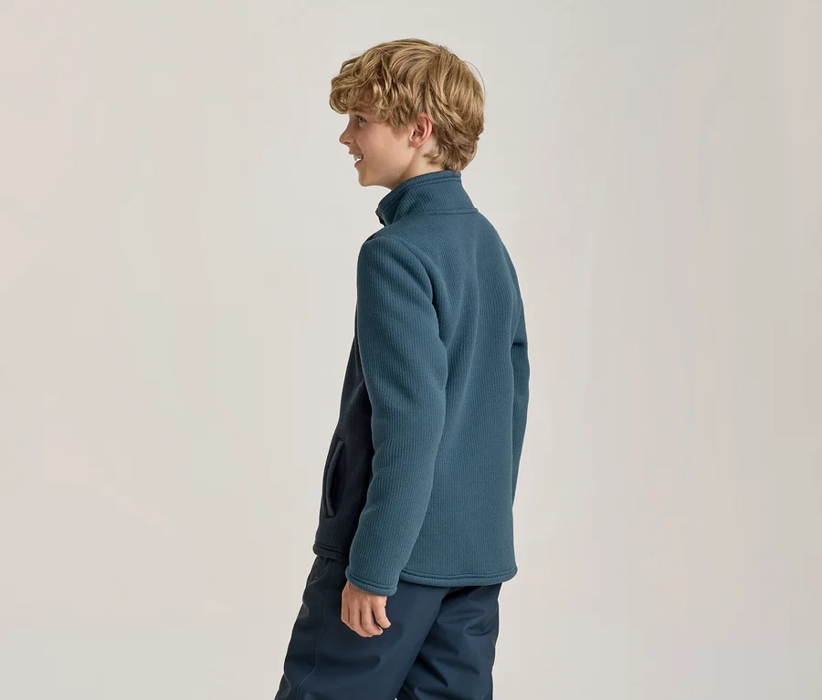 Junger Junge mit blonden Locken, im Profil lächelnd, trägt eine gerippte Fleecejacke in Petrolblau und dunkelblaue Hosen.