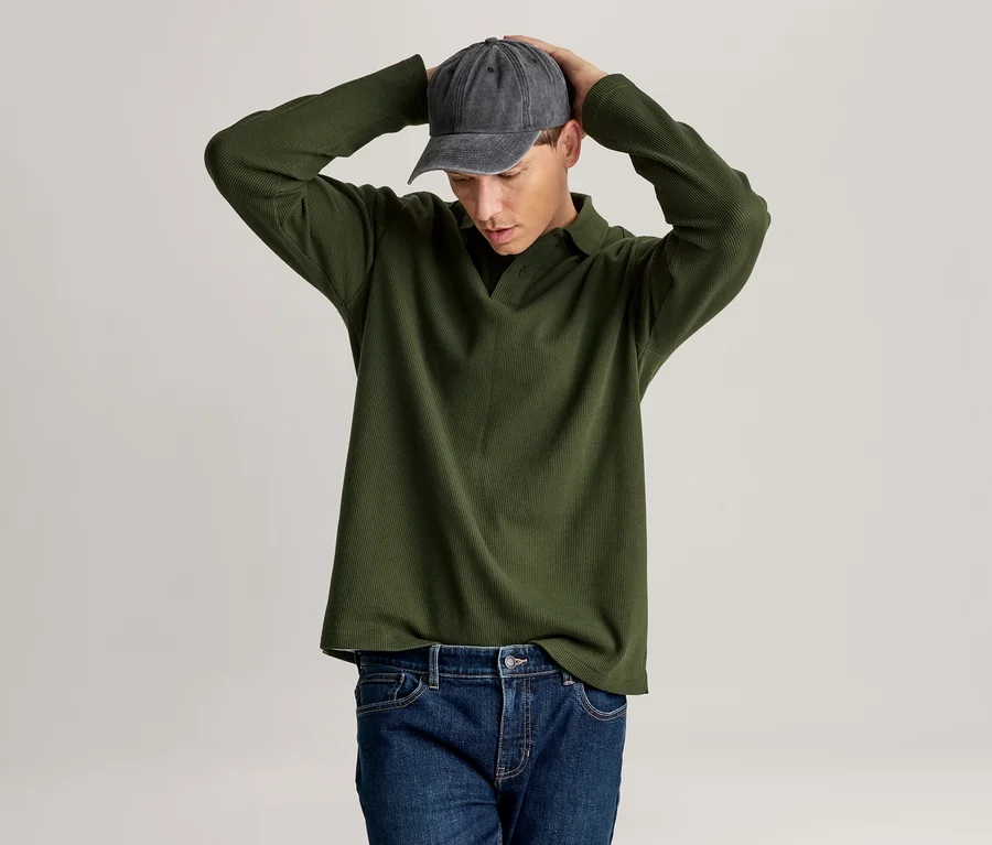 Mann in grünem Langarm-Poloshirt, blauer Jeans und dunkelgrauer Baseballkappe, der mit den Händen am Kopf nach unten blickt.