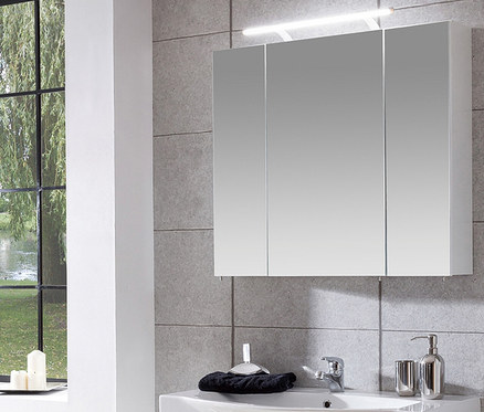 Badezimmer mit einem 80 cm breiten Spiegelschrank "Dorina" mit Licht über einem weißen Waschbecken. Der Schrank hat drei Spiegeltüren. Links ein Fenster mit Blick auf Bäume und Wasser.