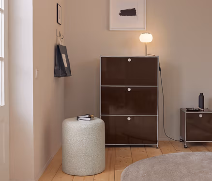 Moderner Eingangsbereich mit hellen Wänden, Holzboden und einem runden beigen Teppich. Ein hoher, dunkelbrauner Modulschrank mit einer leuchtenden Lampe steht neben einem kleineren Rollschrank. Ein heller zylindrischer Hocker mit Notizbuch ist im Vordergrund. Eine gestreifte Einkaufstasche hängt an Haken an der linken Wand.