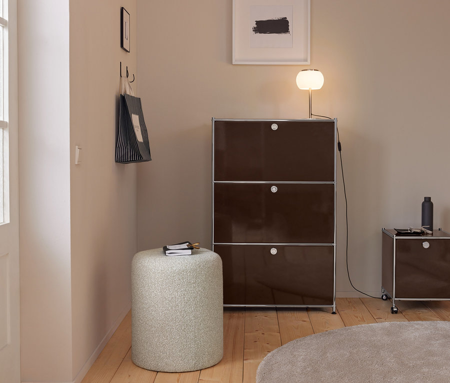 Moderner Eingangsbereich mit hellen Wänden, Holzboden und einem runden beigen Teppich. Ein hoher, dunkelbrauner Modulschrank mit einer leuchtenden Lampe steht neben einem kleineren Rollschrank. Ein heller zylindrischer Hocker mit Notizbuch ist im Vordergrund. Eine gestreifte Einkaufstasche hängt an Haken an der linken Wand.