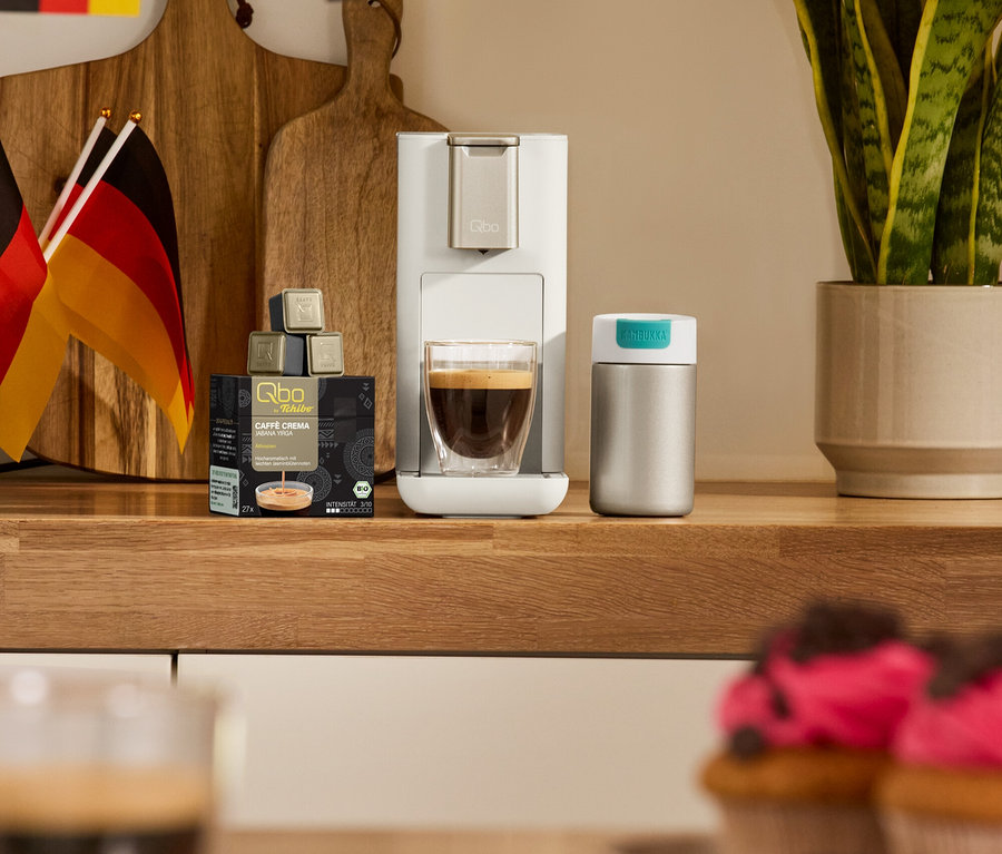 Eine Qbo ICONIC Light Gold Kaffeemaschine mit 2 doppelwandigen Caffè Crema Gläsern und einem Kambukka To-Go-Becher, Light Gold auf einer Küchentheke.