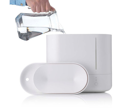 Hand gießt Wasser aus einem Glaskrug in einen weißen Luftbefeuchter.