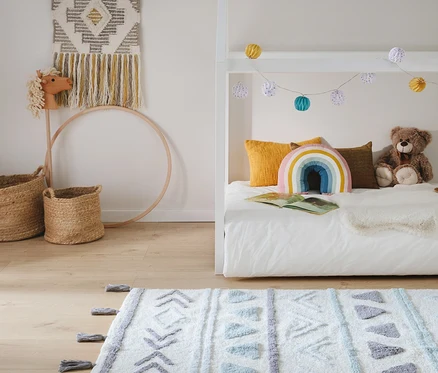 Kinderzimmer mit weißem Bett, gelben Kissen, Regenbogenkissen und Teddybär. Über dem Bett hängen bunte Lichterketten. Auf dem Holzfußboden liegt ein weißer Teppich mit geometrischen Mustern. An der Wand hängt ein Wandteppich, daneben stehen ein Steckenpferd und Körbe.