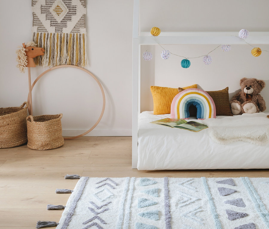 Kinderzimmer mit weißem Bett, gelben Kissen, Regenbogenkissen und Teddybär. Über dem Bett hängen bunte Lichterketten. Auf dem Holzfußboden liegt ein weißer Teppich mit geometrischen Mustern. An der Wand hängt ein Wandteppich, daneben stehen ein Steckenpferd und Körbe.
