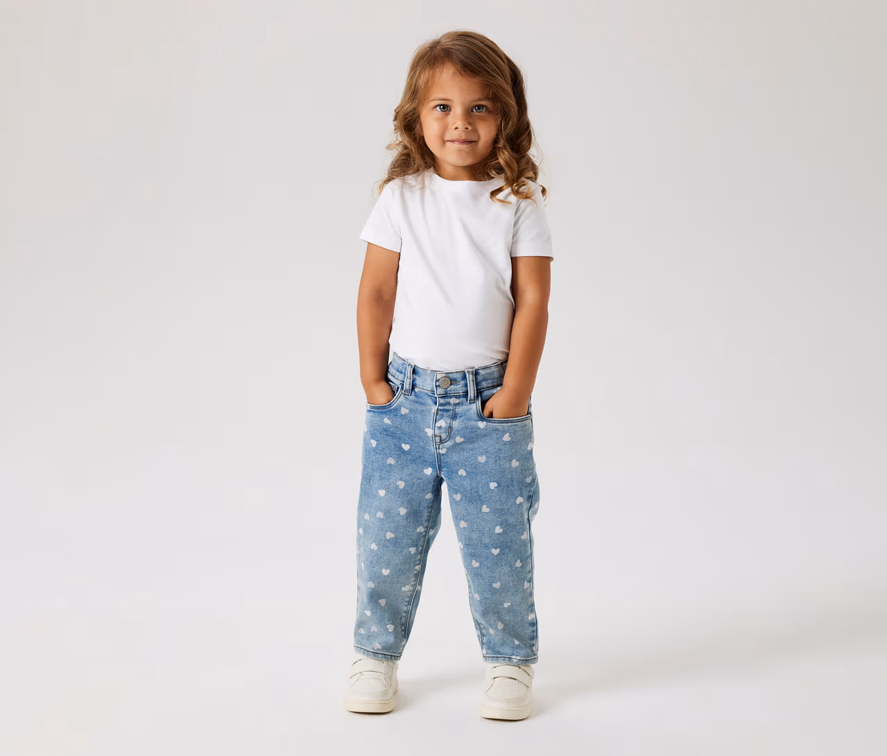 Ein kleines Kind mit langen, welligen braunen Haaren steht frontal, trägt ein weißes T-Shirt, hellblaue Jeans mit weißen Herzmustern und weiße Turnschuhe. Die Hände stecken in den Hosentaschen.