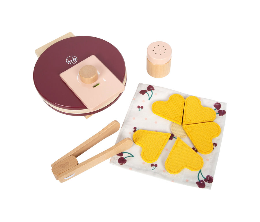 Ein rotes small foot Waffeleisen-Set &raquo;tasty&laquo; mit einem Holzstreuer, einer Holzzange und vier gelben herzförmigen Spielwaffeln auf einer Kirschmuster-Serviette.