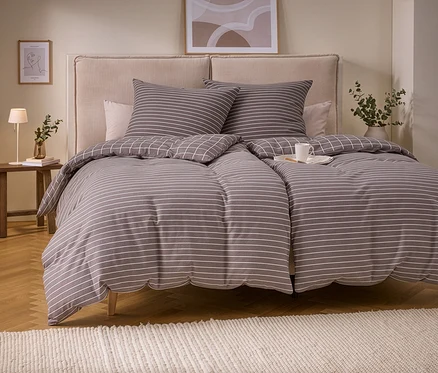 Ein Bett mit Biber-Bettwäsche in Grau mit weißen Streifen und einer karierten Rückseite. Zwei Kissen passen zum Streifenmuster. Eine weiße Tasse steht auf der Bettdecke. Links ein Holzhocker mit Lampe und Vase, davor ein heller Teppich.