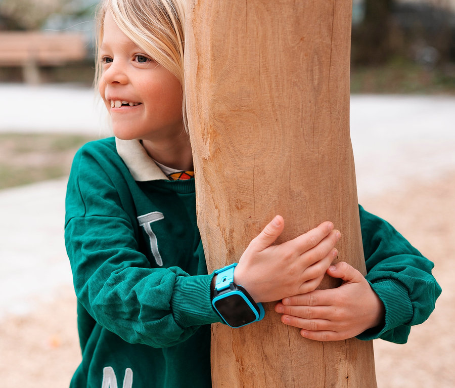 Kind mit blonden Haaren und blauer Xplora XGO3 Kids Watch lugt lächelnd hinter einem Holzpfosten hervor.