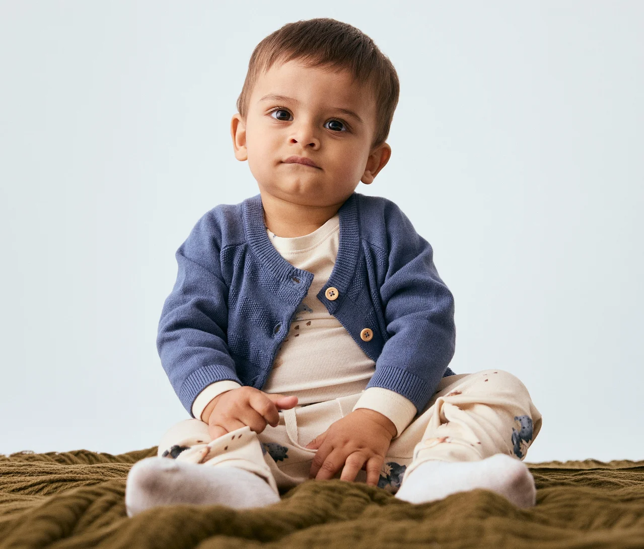 Ein Baby sitzt auf einer Decke und trägt eine blaue Strickjacke, ein hellbeiges Oberteil, gemusterte Hosen und weiße Socken. Es blickt in die Kamera.