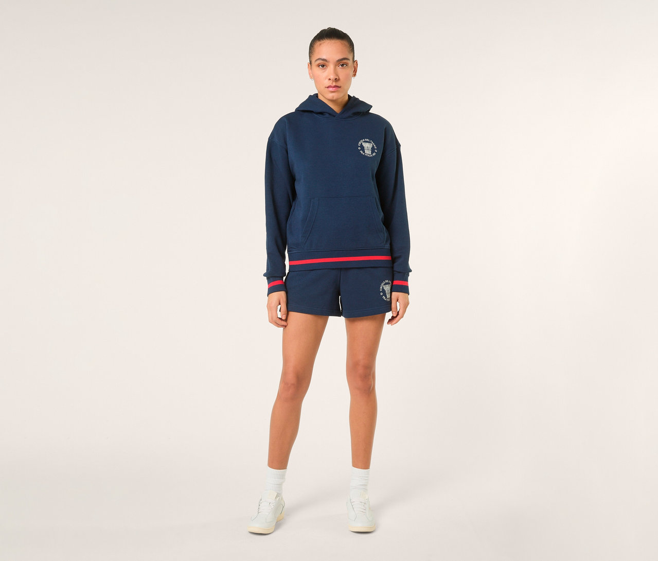Eine Frau steht und trägt einen dunkelblauen FILA Damen-Hoodie »MARLIA« mit weißem Logo und roten Streifen an Ärmeln und Saum, dazu passende dunkelblaue Shorts mit weißem Logo, weiße Socken und weiße Sneaker.