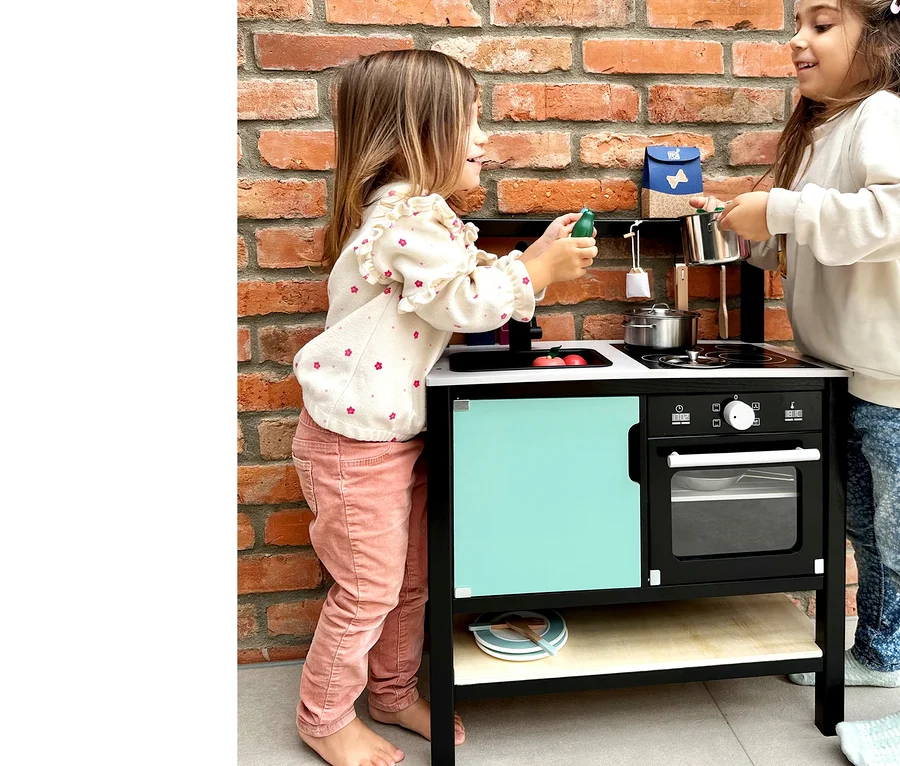 Zwei Mädchen spielen mit der small foot Kinderküche »Industrial« vor einer Ziegelwand.
