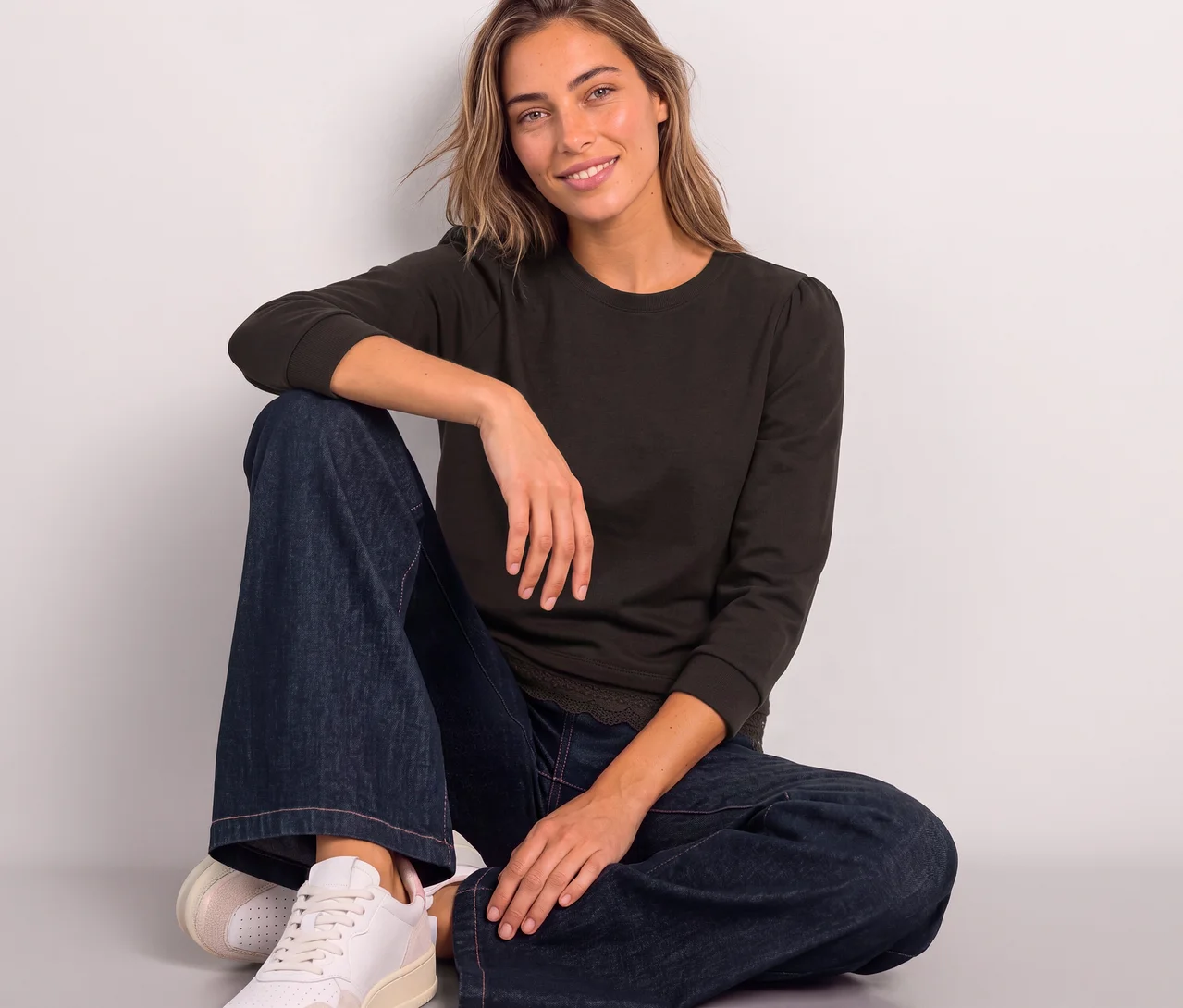 Eine lächelnde Frau sitzt auf dem Boden und trägt ein braunes Street One 3/4-Jerseyshirt mit leicht gepufften Ärmeln, weite Jeans und helle Sneaker.