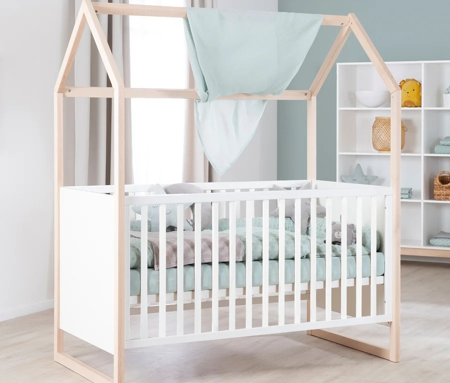 Weißes Babybett mit Holzrahmen in Hausform, hellgrüner Bettwäsche und Stofftieren. Eine hellblaue Decke hängt über dem Rahmen. Im Hintergrund ein weißes Regal mit Spielzeug.