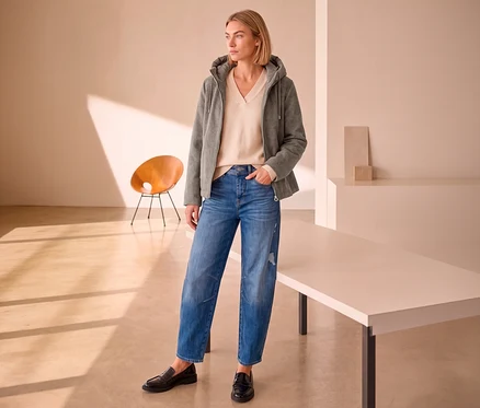 Eine Frau trägt eine graue Street One Übergangsjacke, einen beigen Pullover, blaue Jeans und schwarze Loafer, stehend in einem minimalistischen Raum.