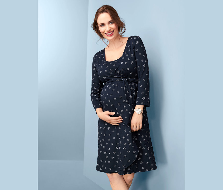 Eine lächelnde, schwangere Frau hält ihren Bauch und trägt ein marineblaues Umstandskleid in Wickeloptik mit Print.