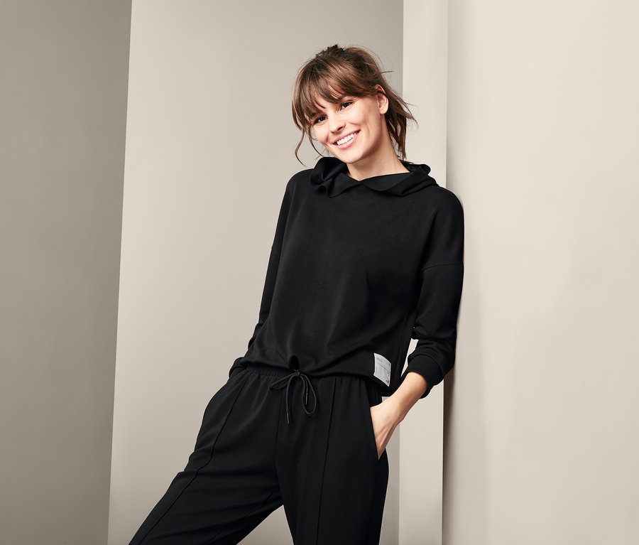 Eine lächelnde Frau lehnt an einer Wand und trägt einen schwarzen Lounge-Sweater und eine Hose.