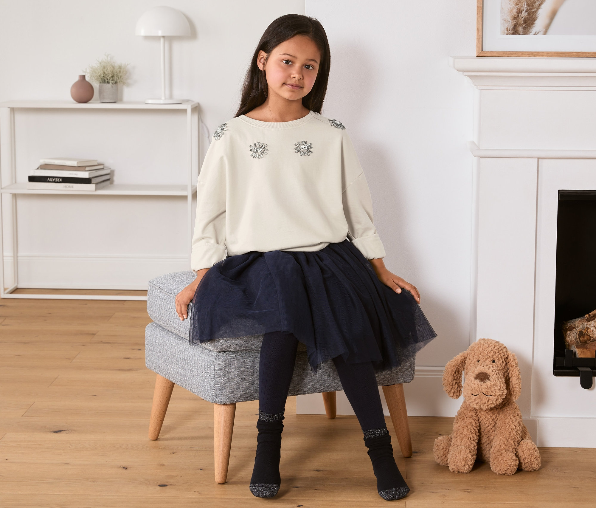 Mädchen sitzt auf Hocker und trägt Sweatshirt mit Strassblumen, Rock und Leggings.