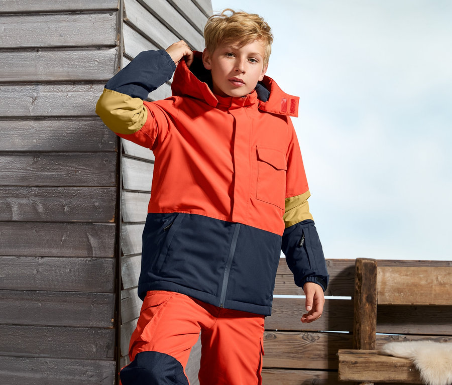 Ein Junge in einer orange-dunkelblauen Snowboardjacke.