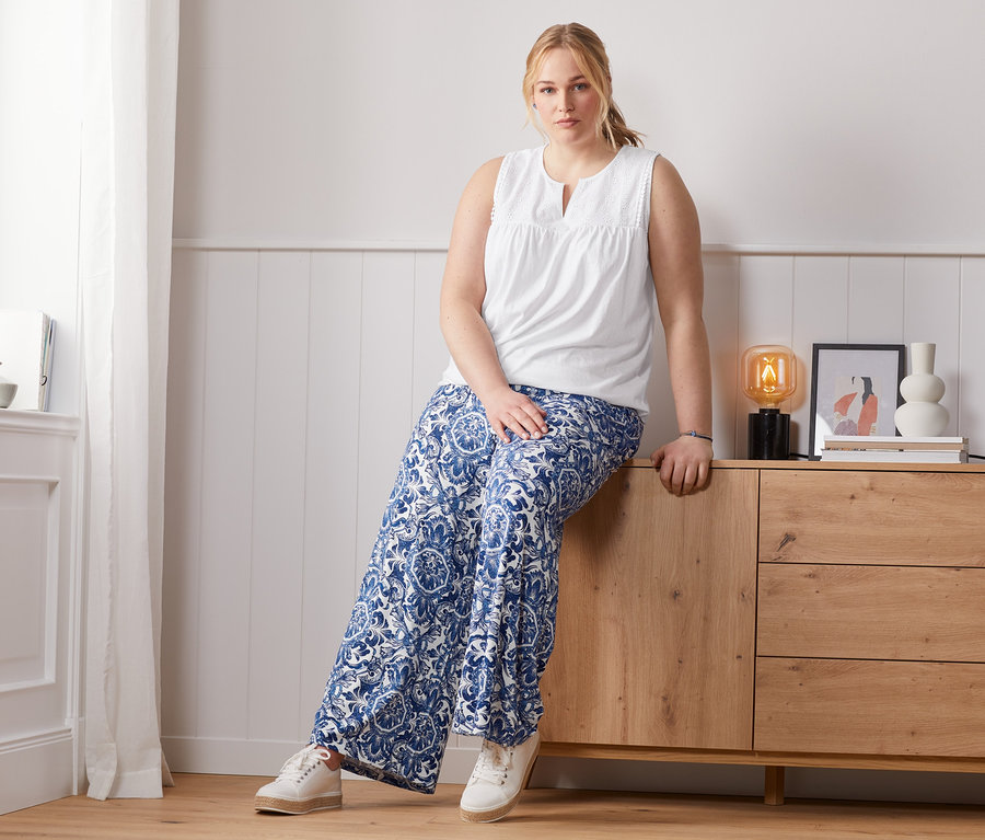 Frau sitzt auf Kommode, trägt bedruckte Palazzohose und Top mit Stickerei.