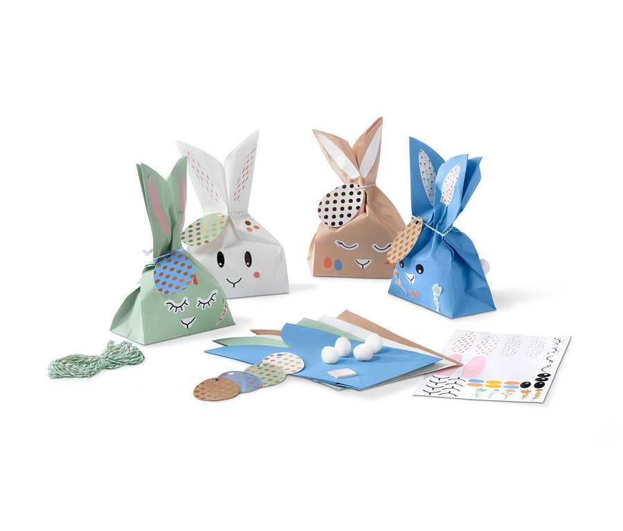 Vier Osterhasen-Geschenktüten mit Gesicht und Ohren stehen nebeneinander. Daneben liegen Dekorationsmaterialien.