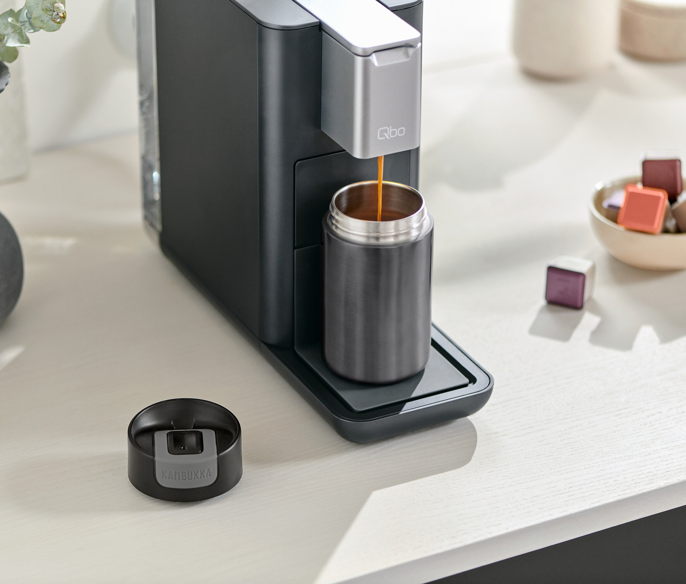 Kaffeemaschine füllt grauen Kambukka To-Go-Becher. Der Deckel des Bechers liegt neben der Kaffeemaschine. Eine Schale mit Kapseln steht daneben.