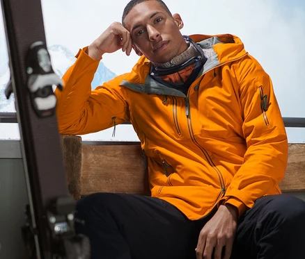 Mann sitzt mit einer orangefarbenen Hardshell-Skijacke auf einer Bank.