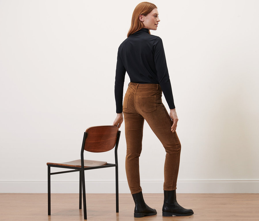 Eine Frau steht seitlich und trägt eine Samthose in Cognac, schwarze Stiefel und einen Rollkragenpullover.