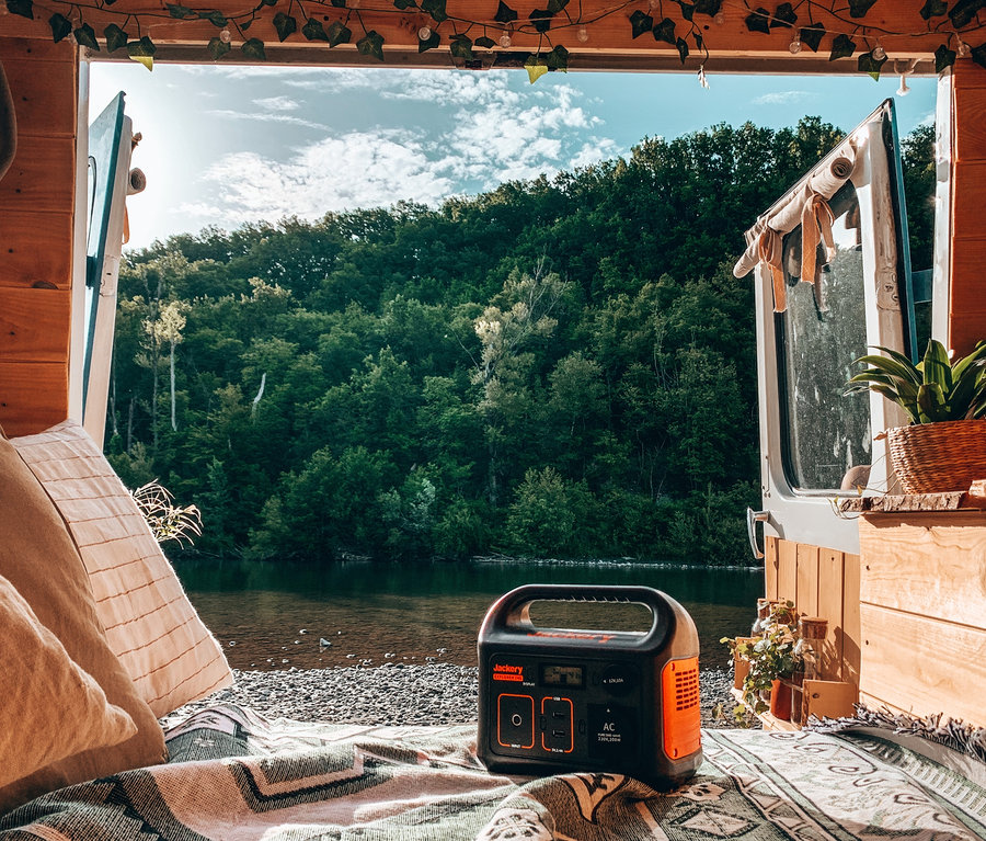 Blick aus dem Inneren eines Lieferwagens auf ein Flussufer mit Wald. Auf einer Decke liegt ein Jackery Explorer 240.