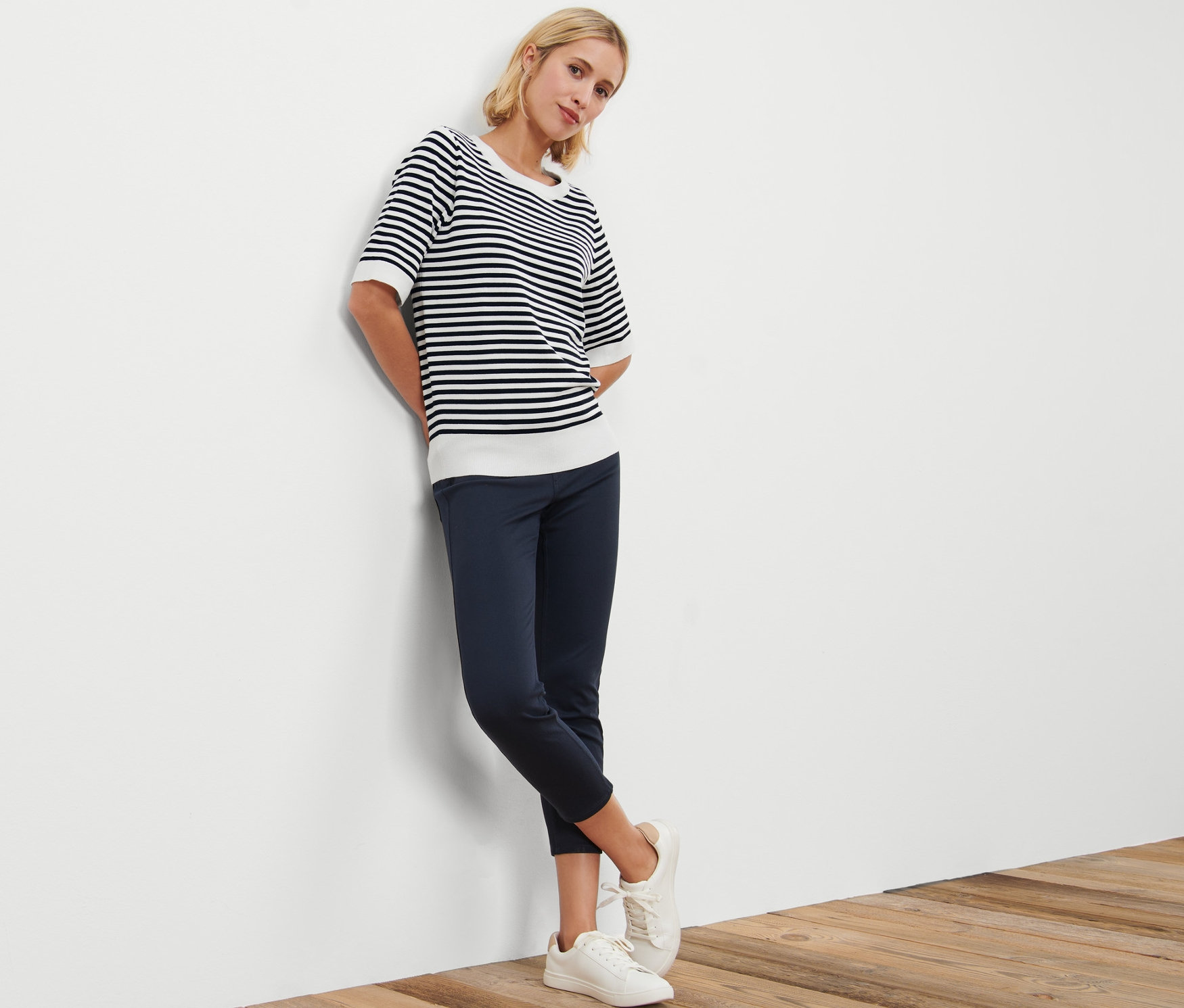 Frau lehnt an Wand, trägt Gestreiften Feinstrickpullover, dunkelblaue Hose und weiße Turnschuhe.