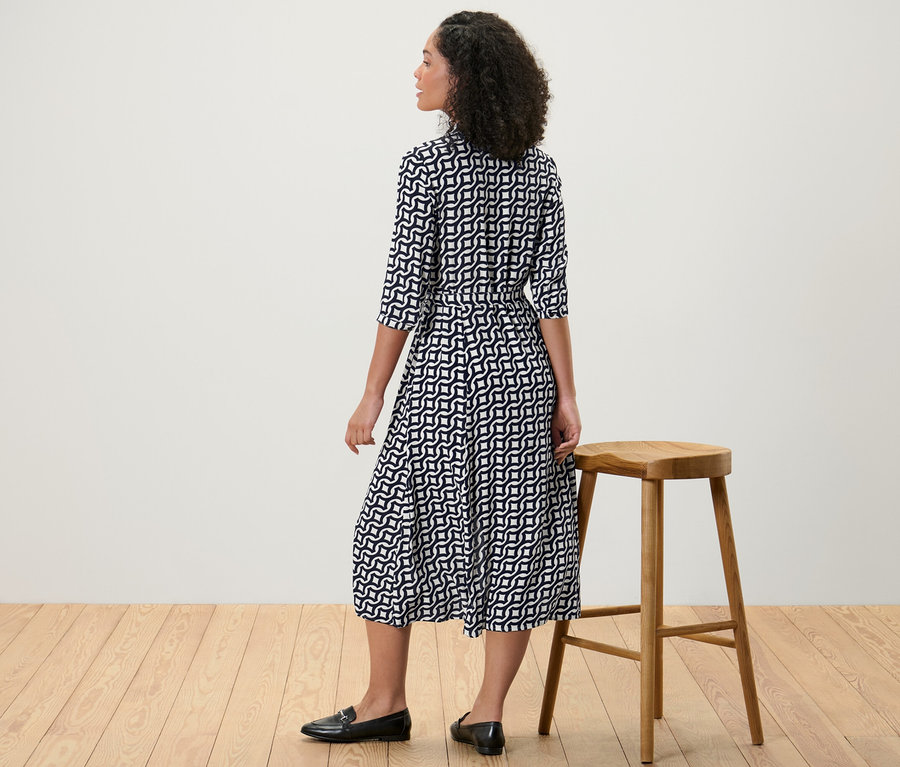 Eine Frau steht neben einem Holzhocker und trägt ein bedrucktes Hemdblusenkleid.