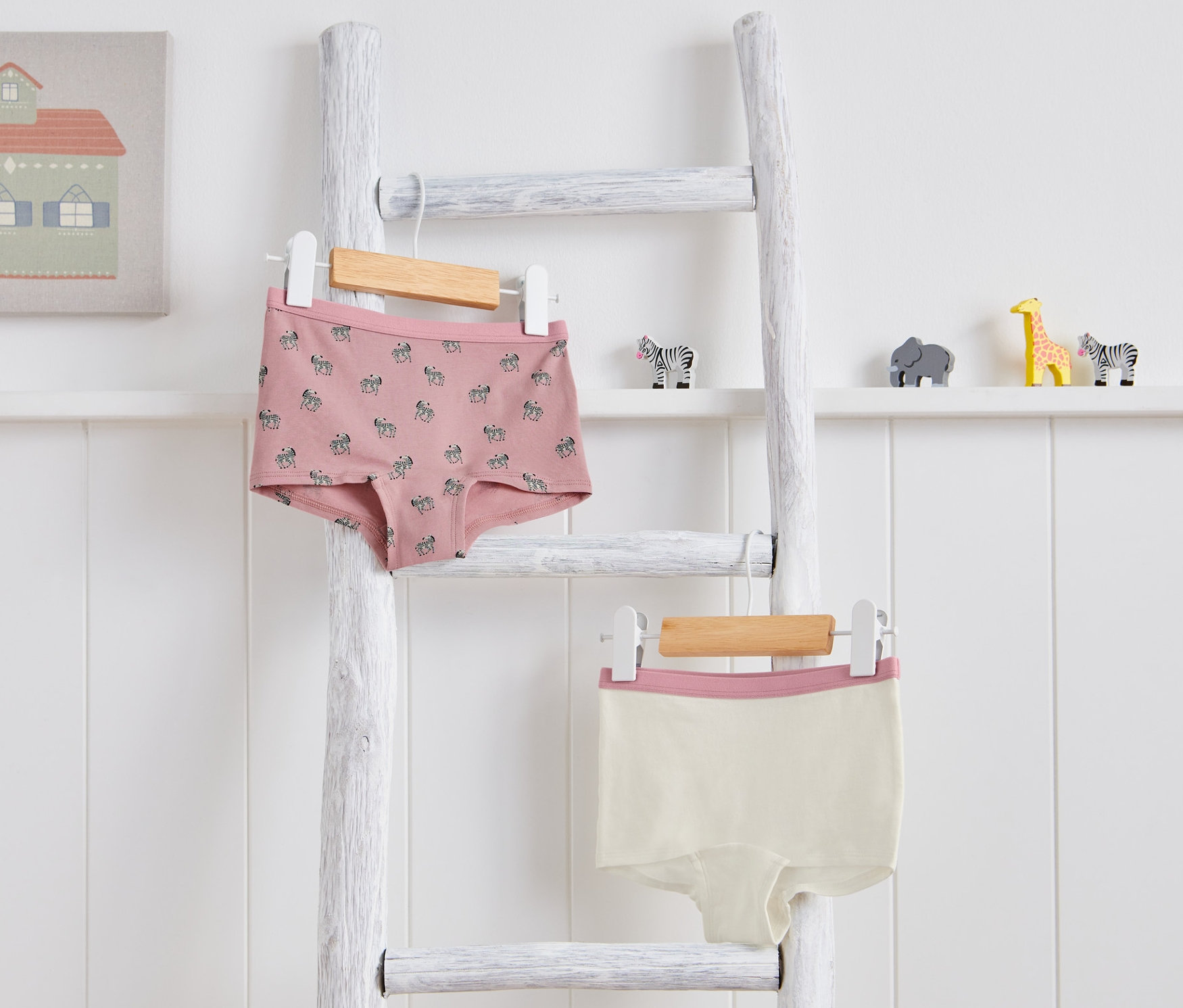 Zwei Kinder-Pantys hängen auf einer weißen Leiter. Eine ist rosa mit Zebra-Muster, die andere cremefarben mit rosa Rand.